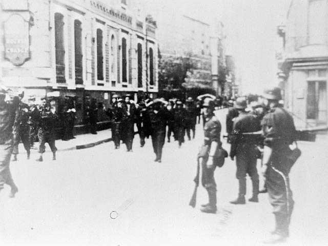 Black and white photograph. A group of Canadian soldiers are marched through the streets of Dieppe; their hands are behind their heads. German soldiers stand to the side, armed, watching them.