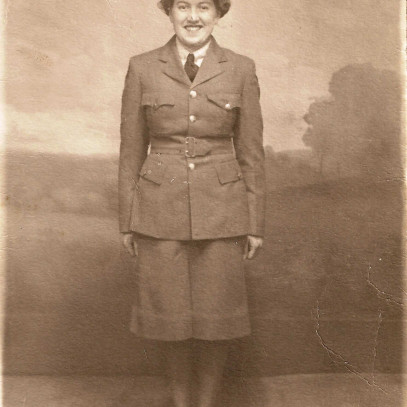 Sepia tone photo. A young women is shown in full military dress, in a skirt uniform. She is grinning standing at attention.