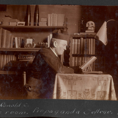 Black and white photograph. RC MacGillivray is shown from the side, sitting at a table covered in a long, embroidered cloth, reading from a book. There are book shelves behind him. He is wearing a dark uniform and a hat.