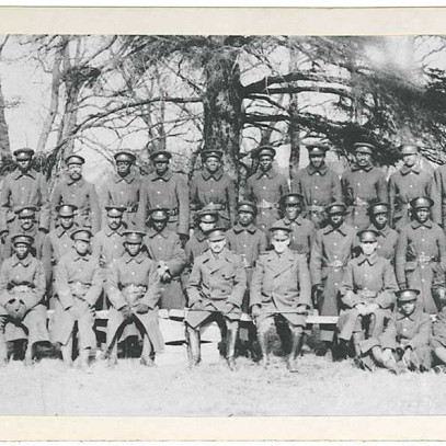 Black and white photograph. A large group of majority black men sit for a group photo. Most are wearing long military coats. Trees and rooftops are visible in the background.