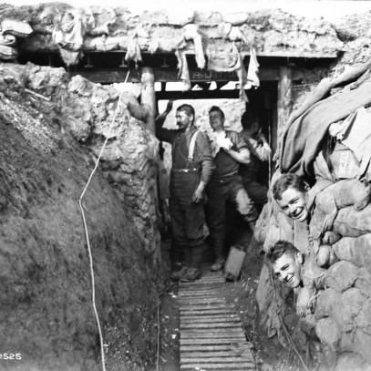 Black and white photograph. A trench with a wooden walkway. Two men stand underneath a wooden "bridge". Others peek out from bunk holes built into the sandbagged wall.