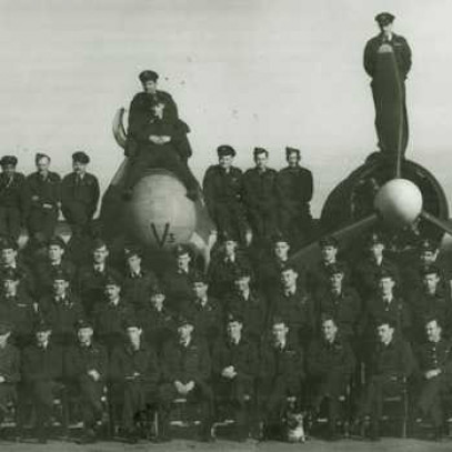 Black and white photograph. About 60 members of 404 squadron pose for a group shot in front of a Beaufighter. Men are lined up in front of the plane and also sitting on the engines and standing on the wings.