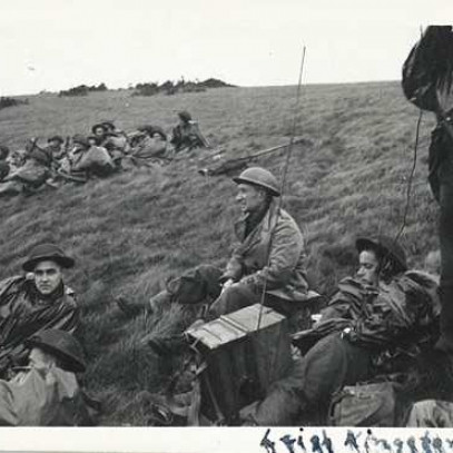 Black and white photograph. Archie, in full gear, sits on a grassy hill, surrounded by other lounging soldiers. He's looking away from the camera. There's blurred writing on the corner of the photo.