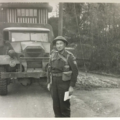 Black and white photograph. Archie stands at attention, with a smile, in front of a military vehicle.