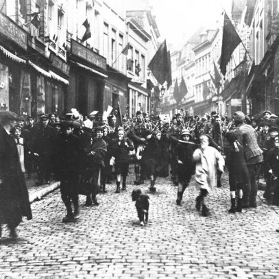 Black and white photograph. 42nd Battalion, playing bagpipes, marches a crowd of French citizens the streets of Mons.