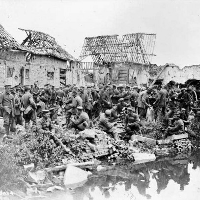 Black and white photograph. A large group of German prisoners of war gather along the side of a river. Rubble and shrubs litter the ground. Ruins surround the men.