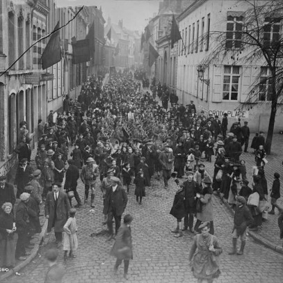 Black and white photograph. A group of Canadian soldiers, playing bag pipes, marches through a crowded, cobblestoned street. Buildings line the street, and the group appears to be approaching an open area.  French flags hang from buildings.