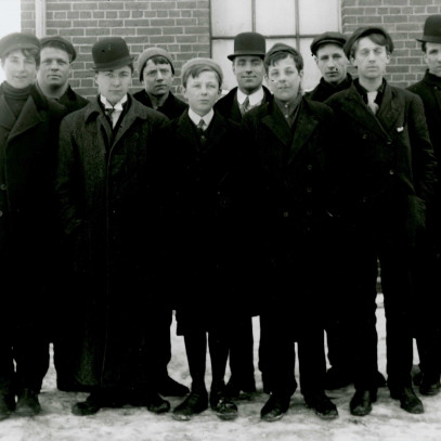 Black and white photograph. A group of ten men stand together on the snow covered ground in civilian clothing; most faces show signs of excitement.