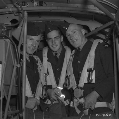 Black and white photograph. Three airman, holding bags (parachutes?) and wearing straps stand inside an aircraft, surrounded by instruments and metal piping. They all crouch a little, the ceiling is low.