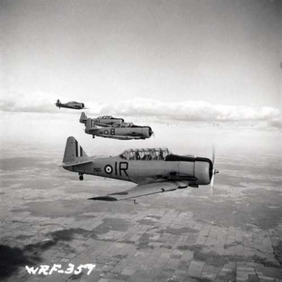Black and white photograph. Four aircraft fly through the air in a line beside each other. On the ground, the patchwork appearance of fields.