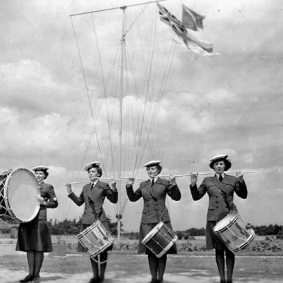 Black and white photograph. 4 women in navy uniforms stand in a line. The furthest left plays a large base drum. The other three have snare drums, their sticks held horizontally in the air. The Red Ensign flies behind them.