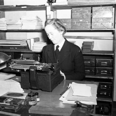 Black and white photograph. A woman with styled hair sits at a desk, typing on a typewriter. Behind her are shelves filled with paperwork and boxes. There are additional papers on the desk, as well as a small lamp.
