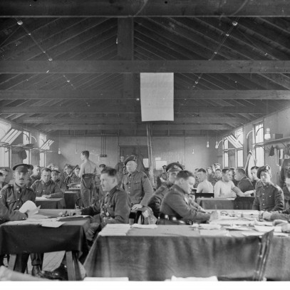 Black and white photograph. Men, some in full uniform and some without shirts, sit around tables in a large room. The windows are open wide to let in light and air. Some of the men look at the camera.