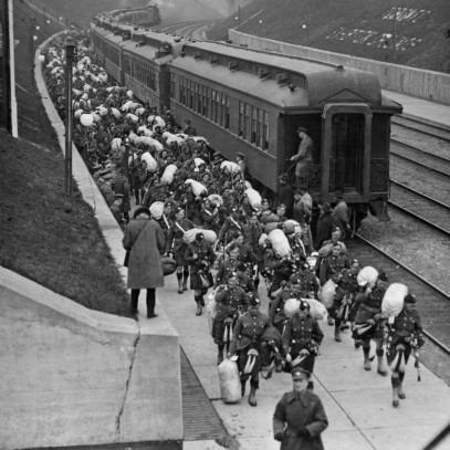Black and white photograph. Hundreds of men in military kilts walk beside a train, disembarking. The tracks run in between two grass hills ending in a cement wall; a reporter stands on the wall taking notes or a photograph.