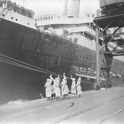 Black and white photograph. Women wave handkerchiefs on a dock while men board a ship.