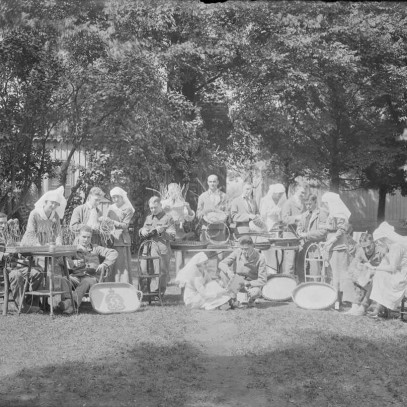 Black and white photograph. Nurses and soldiers sit on chairs or on the ground near a large tree. They ware weaving baskets.