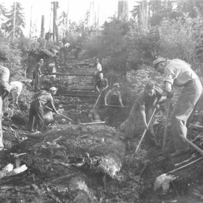 Black and white photograph. Men are clearing a path through a forest to build a road.