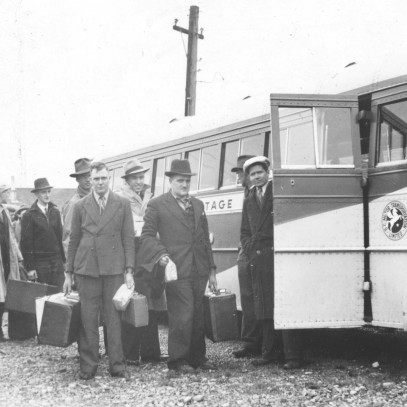 Black and white photograph. Well-dressed men congregate in front of a large bus, holding suitcases and waiting to board. Most are facing the camera. A second bus is to their other side.