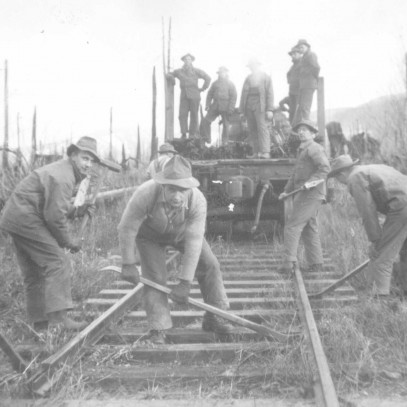 Black and white photograph. 5 men build or repair railway tracks using long handled tools. 5 other men stand watching on the back of a pick-up truck. They are in a forested area.