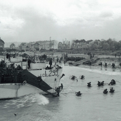 Black and white photograph. A large boat is anchored off the beach. Men wade through waist-high waters, many carrying equipment such as bicycles, toward the beach. Houses line the coast.