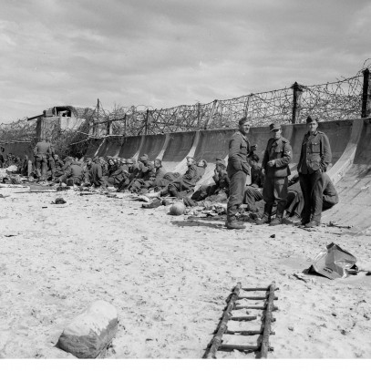 Black and white photograph. A sandy beach meets a high, curved seawall topped with thickly laid barbed wire. A large machine gun emplacement is visible further down the beach. A line of German soldiers, now POWs, sit against the seawall.