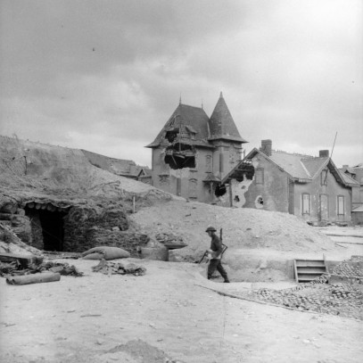 Black and white photograph. Stone buildings are in the background, with large shell holes. A lone Canadian soldier walks into a sandbagged bunker built under a large sand dune.