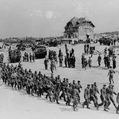 Black and white photograph. A long column of German soldiers marches along the sandy beach. Canadian personnel watch them. Beach obstacles and military vehicles are visible. Canada House looms over the column of POWs.