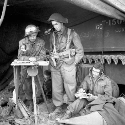 Black and white photograph. Soldiers stand in a make-shift shelter built against a tank.