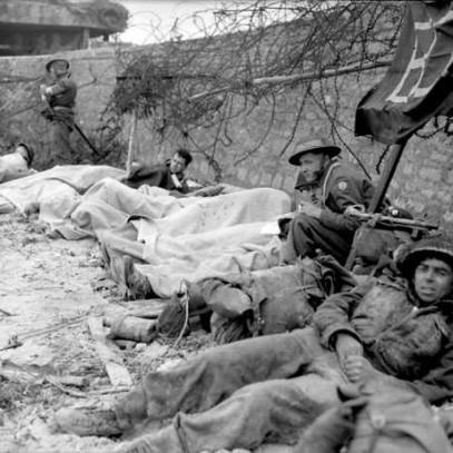 Black and white photograph. Wounded soldiers lie against a stone wall on the beach. Some are completely or partially covered with white sheets, while others sit up on their elbows and look at the camera.
