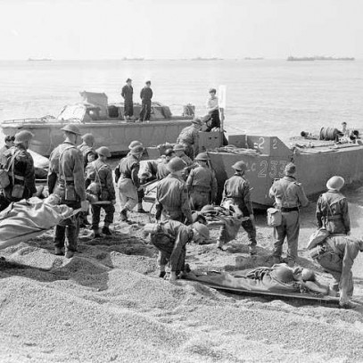 Black and white photograph. On a rocky beach, men in battle dress stand with their backs to the camera looking at the two small boats on the shoreline. Some hold stretchers with men on them. They wait to board the stretchers on the boats.