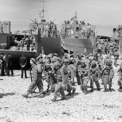 Black and white photograph. A large group of soldiers in battle dress walk onto a rocky beach from landing craft ramps. Navy personnel await the landing of a 2nd craft.
