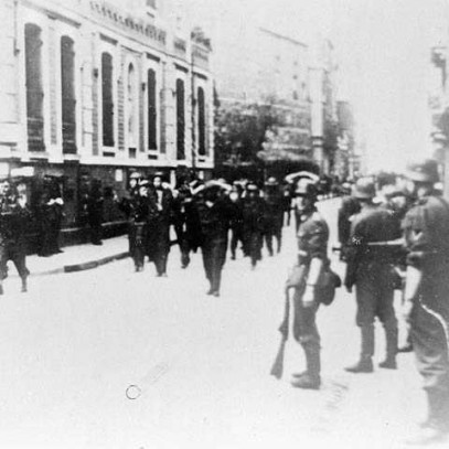 Black and white photograph. A group of Canadian soldiers are marched through the streets of Dieppe; their hands are behind their heads. German soldiers stand to the side, armed, watching them.