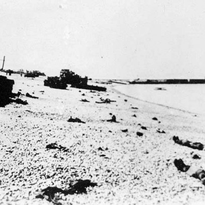 Black and white photograph, a pier visible in the distance. Tanks and bodies lay on the rocky beach.
