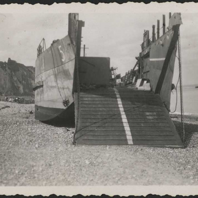 Black and white photograph. A landing craft on the pebbly beaches at Dieppe, landmark white cliffs in background. The craft is fully on the beach, and it's landing platform is down.