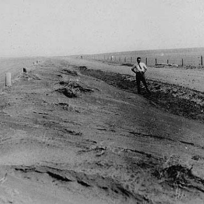 Black and white photograph. A man stands on the side of a road, flanked by fields. Despite the black and white colouring of the photo, it is easy to tell how dry the ground is. Dirt drifts through the air.