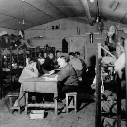 Black and white photograph. A small room with low, curved roof. Bunk beds, many filled with men, line both walls. There are tables in the middle of the room. Men stand around, or sit at the tables.