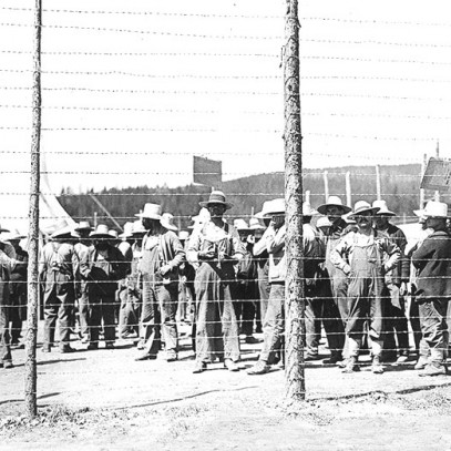 Black and white photograph. A large group of men, dressed in overalls and wide-brimmed hats, stand idly behind a high barbed wire fence. A tent is visible in the background.