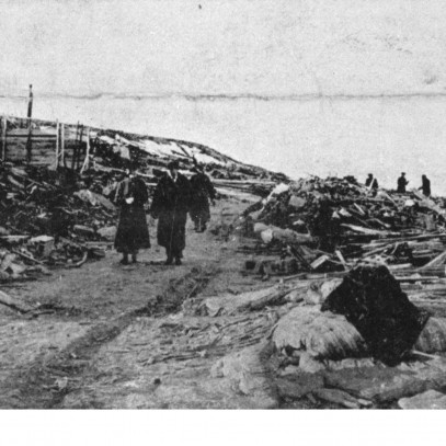 Black and white photograph. Survivors walk along a narrow path through the debris, away from the shoreline. The skeletal remains of a large building are on the left.