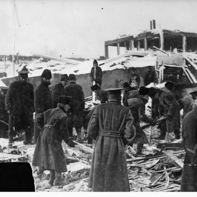 Black and white photograph. A group of soldiers digs through debris, searching for victims of the explosion. They wear long winter coats. The skeletal remains of a building  are visible on the hill beyond them.