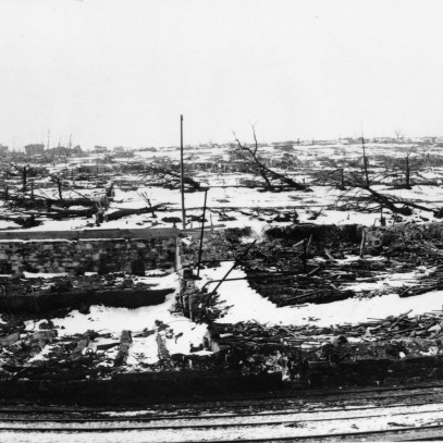 Black and white photograph. Train tracks in immediate foreground. Some structures visible in distance. The foundation and rubble of a building is seen in the centre of the photo. Charred trees and snow on the ground is visible.