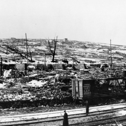 Black and white photograph. Train tracks in the immediate foreground. Snow, rubble, charred trees, and the ruins of buildings can be seen everywhere. Notable, a destroyed rail car is visible.