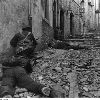 Black and white photograph. Canadian soldiers crouch and aim against a wall on a narrow street. A casualty is visible in front of them.