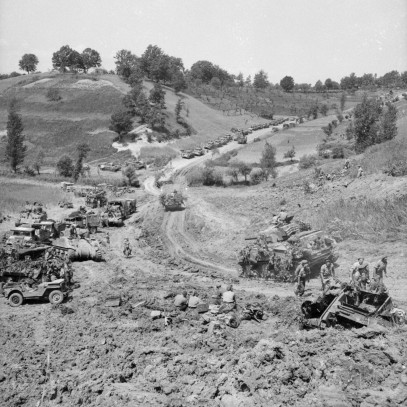 Black and white photograph. A dirt road cuts through the hills, covered sparsely with trees. At the edge of the road, Canadian soldiers investigate an overturned vehicle. Other military vehicles line the road.