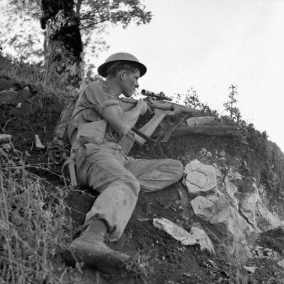 Black and white photograph. A Canadian soldier crouches underneath a tree, his body sheltered by a rise in the rocky, brush covered ground. His sleeves are rolled up. He holds  his gun at ready, and his eyes are squinted in concentration.