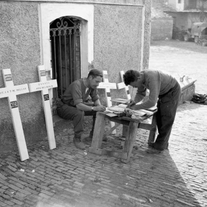 Black and white photograph. 4 white crosses with names lean against a stone wall beside a metal gate. Two men lean over a small wooden table preparing more crosses.
