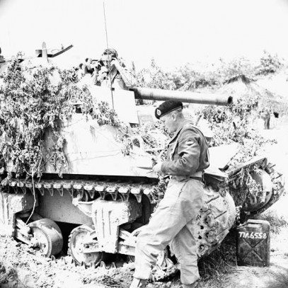 Black and white photograph. A tank is partially covered in branches as camouflage. A soldier peeks out of the top of the tank, and another stands in front. A gas can is on the ground.