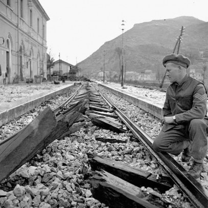 Black and white photograph. A broken rail track runs past a large building and into the mountains. A Canadian kneels beside the rails examining the broken wooden ties, which the camera is focused on. All the ties have been cut in half.