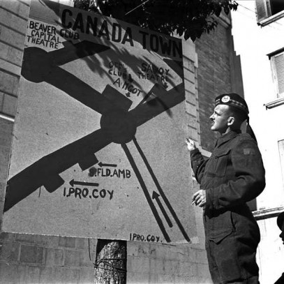 Black and white photograph. A soldier poses in front of a sign reading "Canada Town", which provides directions to various clubs in the area.