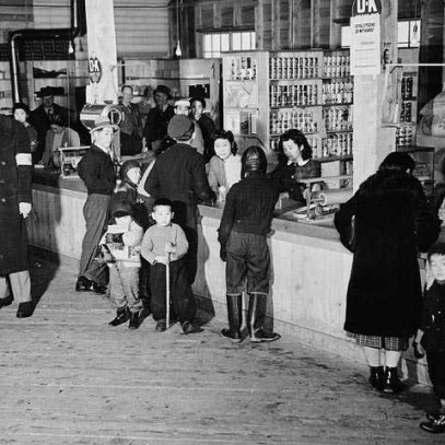 Black and white photograph. People stand in front and behind a store counter, with shelves full of canned food and other stock. People converse across the counter.