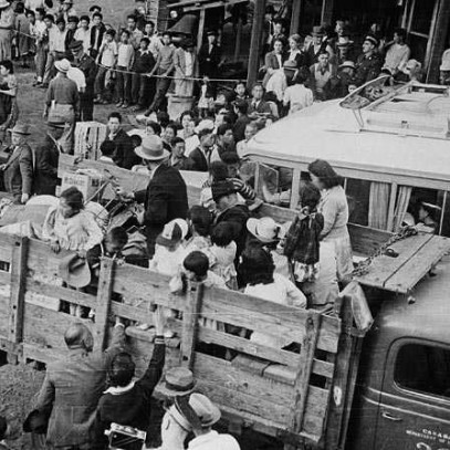 Black and white photograph. Men, women, and children are loaded into the back of an open-bed pick-up truck with their belongings. People reach up to say goodbye. Other families wait their turn.